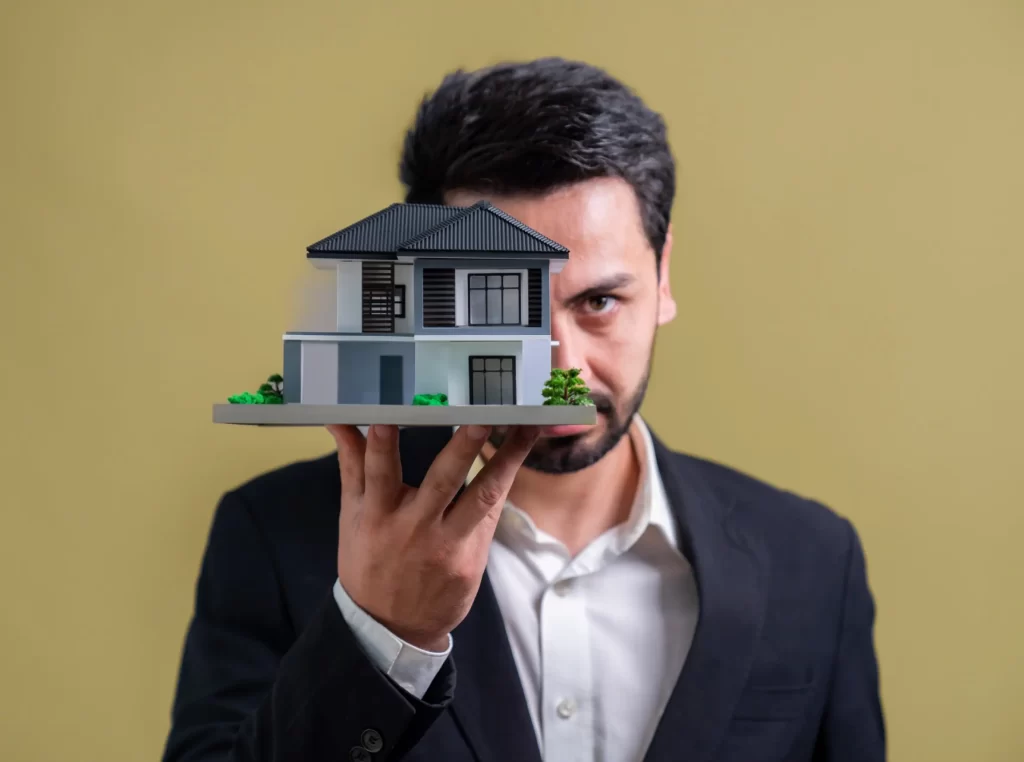 Homme en costume tenant devant lui une maquette de maison moderne, la main levée à hauteur du visage, sur fond uni beige